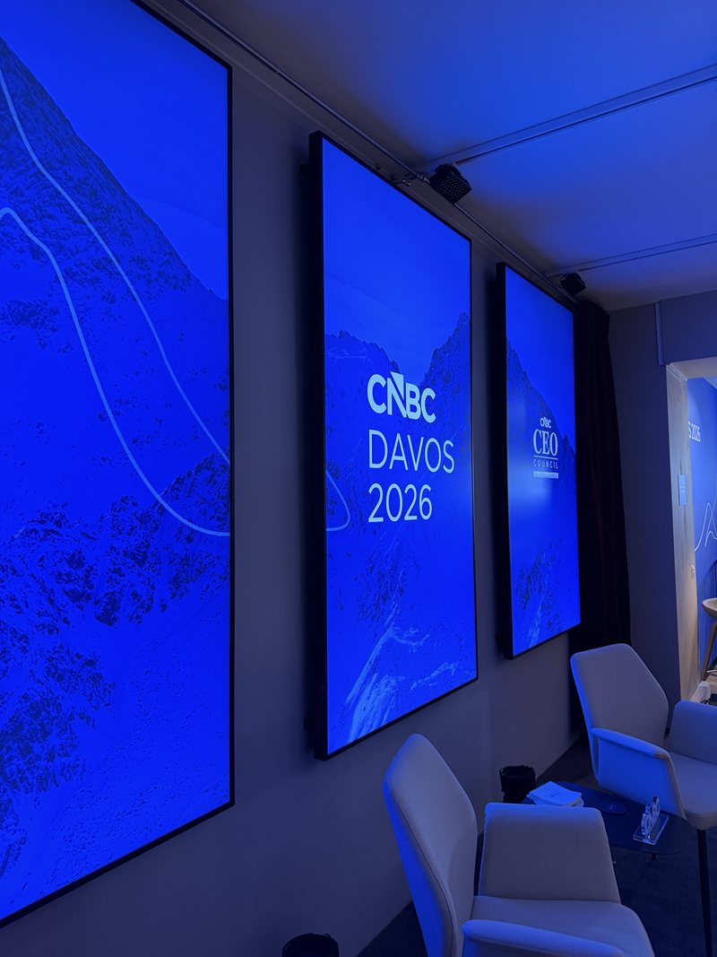 Innenraum des CNBC House in Davos: drei blau leuchtende Bildschirme mit Bergmotiven und dem Schriftzug „CNBC Davos 2026“, davor helle Sessel.