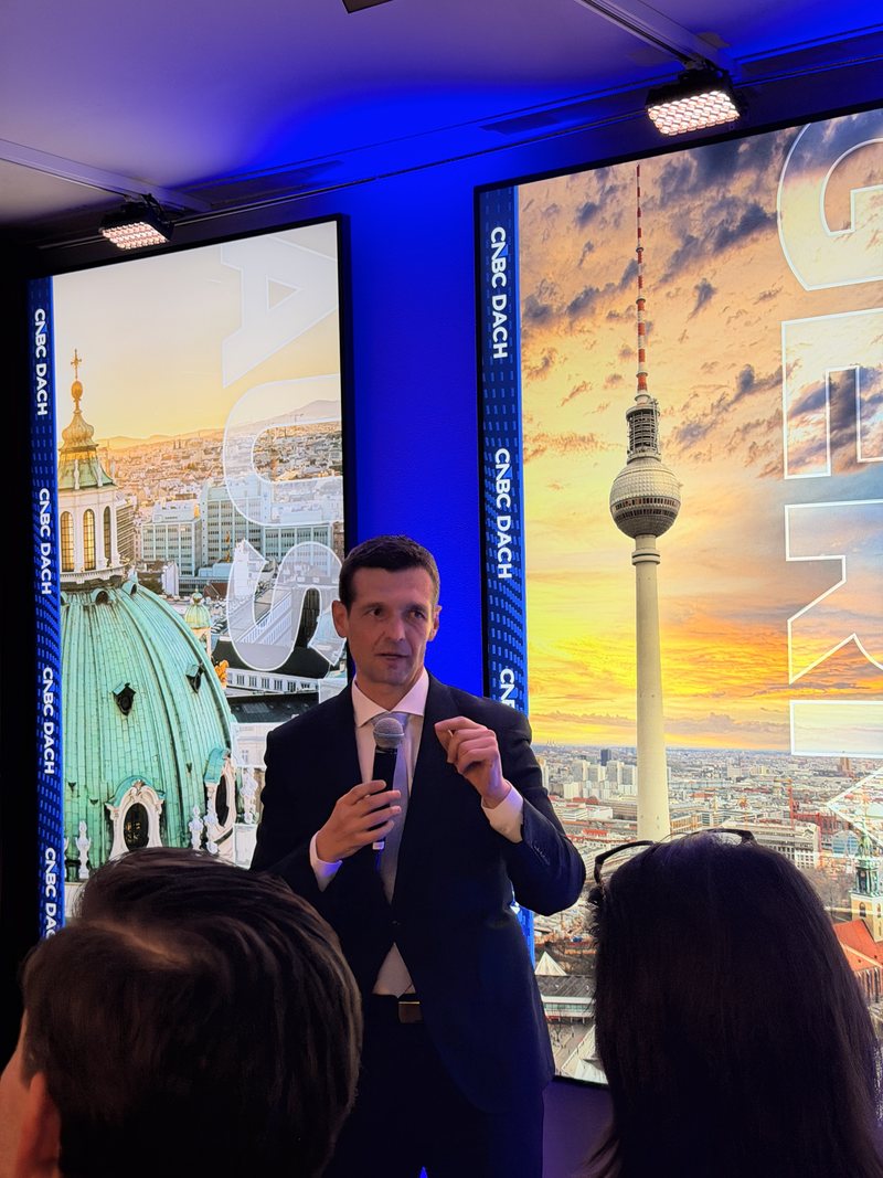 Rusmir Nefić spricht in Davos mit Mikrofon vor Gästen, hinter ihm Bildschirme mit CNBC-DACH-Motiven von Wien und Berlin.